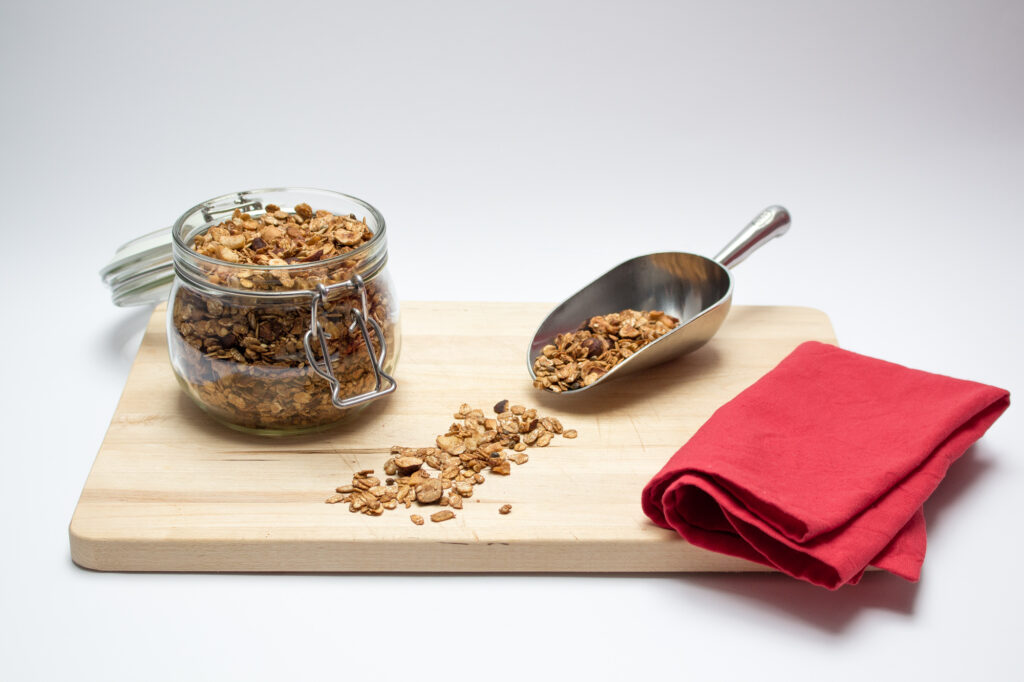 Granola in a jar and on a ladle presented on a wooden board with a red napkin on the side.