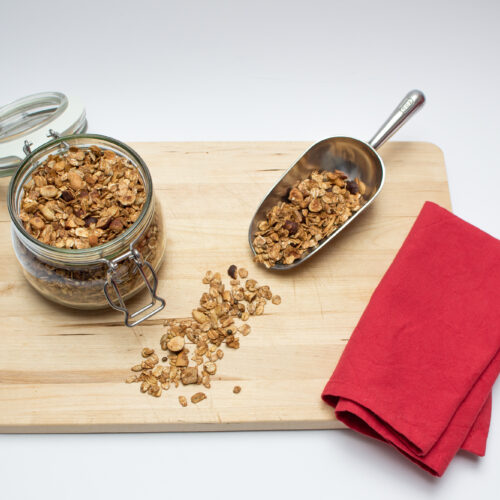 Granola in a jar and on a ladle presented on a wooden board with a red napkin on the side.