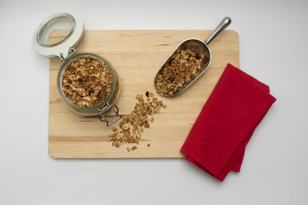 Granola in a jar and on a ladle presented on a wooden board with a red napkin on the side.