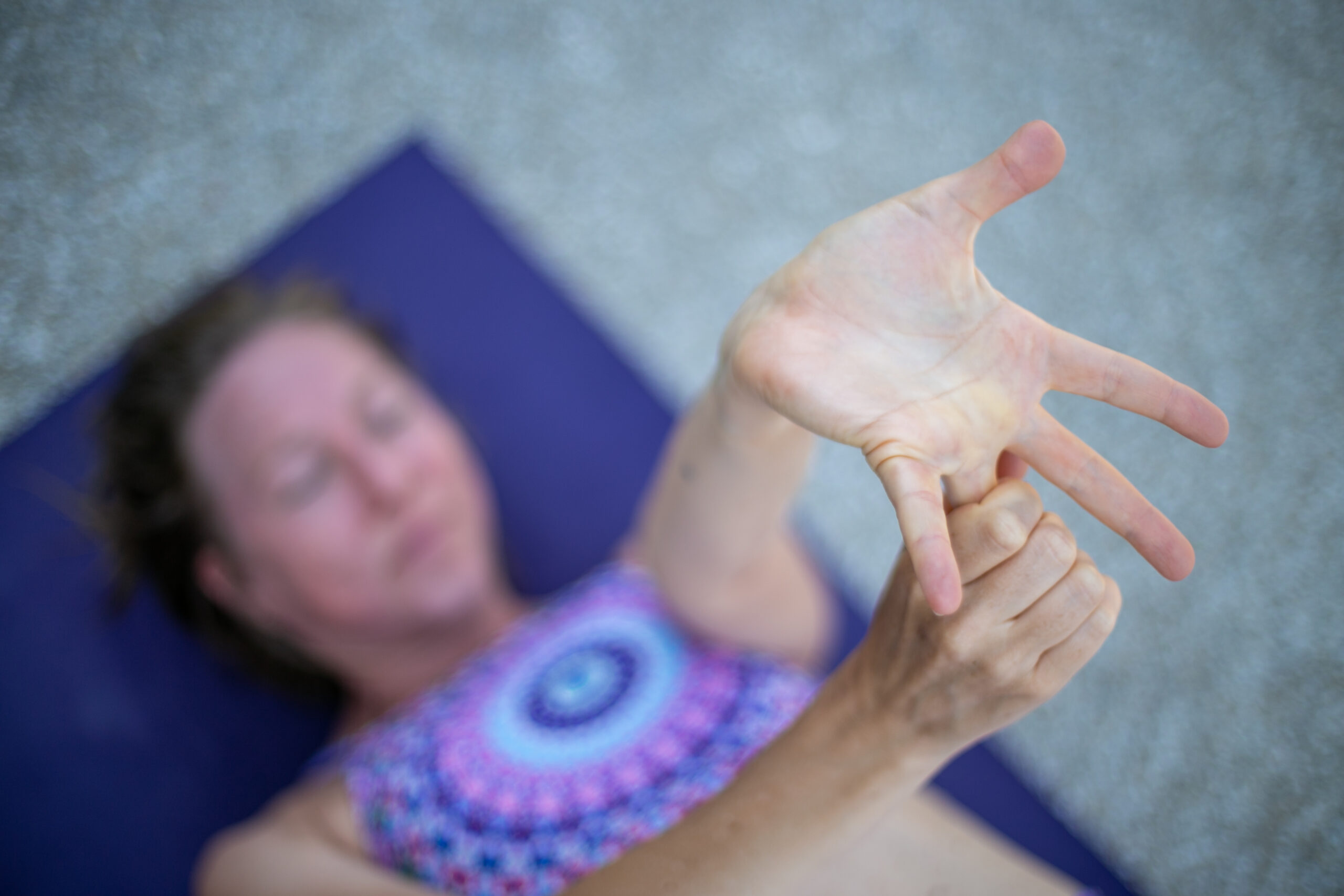 A blonde woman in colourful yoga clothing lying on her back on a purple yoga mat doing wrist stretches.